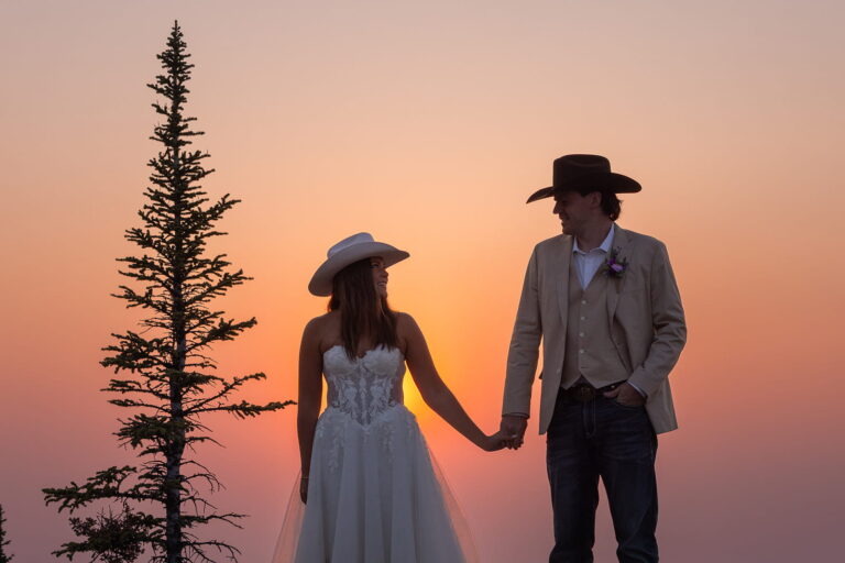 A bride and groom wearing cowboy hats stand next to a pine tree in front of a bright pink sunset sky.