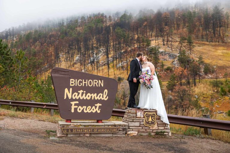 A bride and groom kiss while standing next to the Bighorn National Forest park sign during their Bighorn elopement wedding in Wyoming.