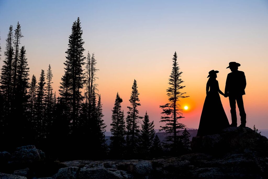 A silhouette image of a bride and groom wearing cowboy hats in front of an epic sunset in the Bighorn National Forest.