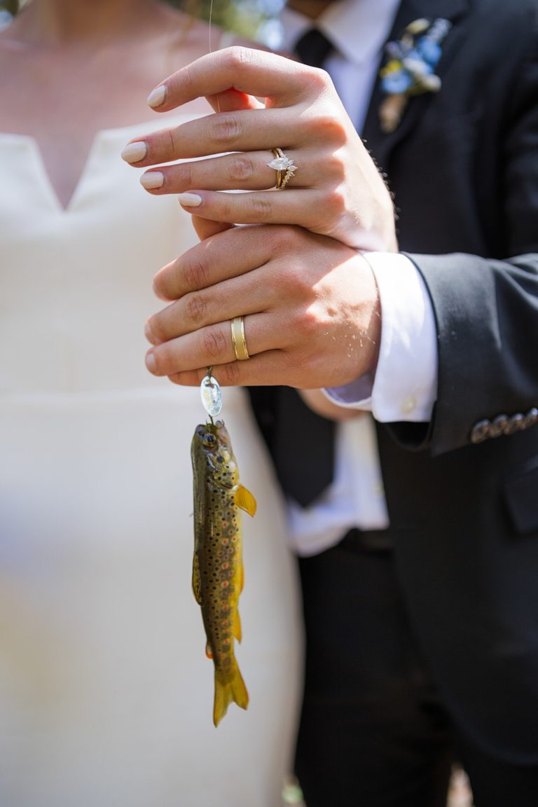 The hands of a married couple with their wedding rings hold a fishing line with a trout on the end of it. In the background of the image you can see the wedding dress and tux of the couple. It's the first fish of the day during their elopement in Bighorn National Forest.