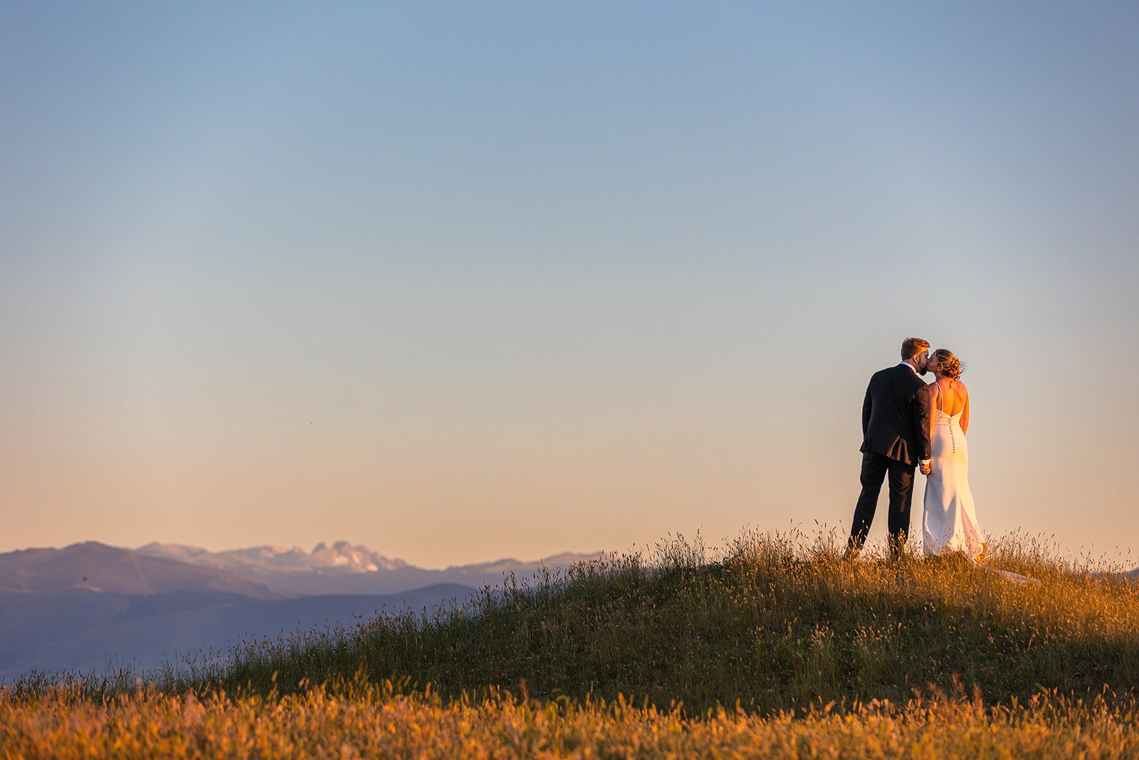 A couple in their wedding clothes kisses on top of a grassy hill outside of Sheridan, Wyoming. In the background, the mountains of the Bighorn National Forest can be seen, glowing purple in the light of the setting sun.