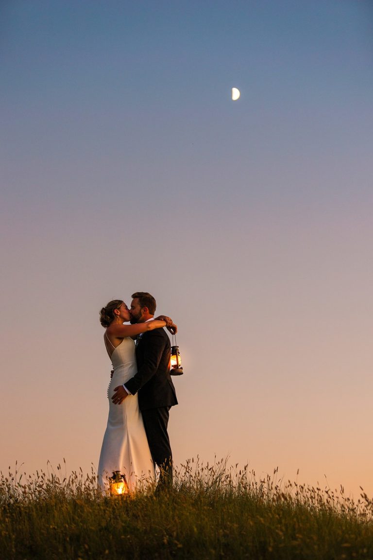 A newly married couple embraces on top of a grassy hill during their elopement in the Bighorn National Forest. The sky is glowing with the colors of sunset and the couple is illuminated by the glow of two lanterns. A sliver of the new moon can be seen in the sky behind them.