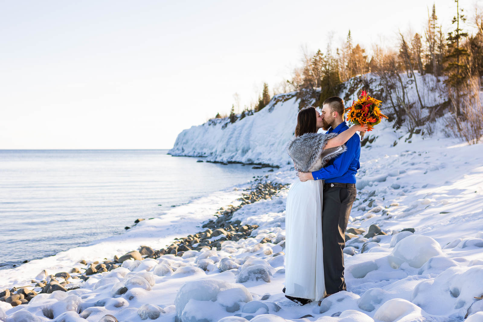 A couple kiss while standing in the snow along the frozen shore of Lake Superior during their winter elopement along the North Shore. Winter is one of the best times of year to elope in Minnesota!