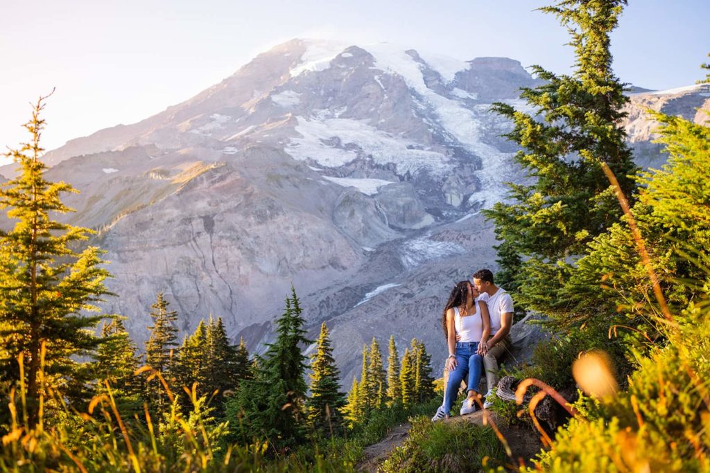 At Mount Rainier National Park, a couple sits below an evergreen tree as the golden light of sunset glows on the great mountain behind them.