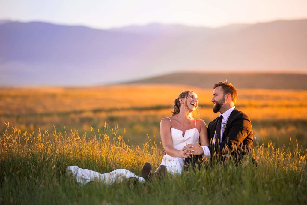 The colors of sunset glow gold and purple around a couple in their wedding clothes laughing while sitting in a grassy field. An epic mountain backdrop behind them makes it clear that they chose a beautiful place to elope.