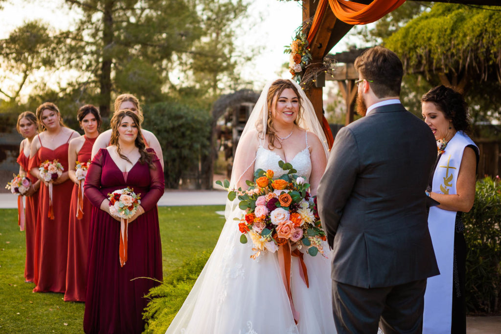 During their wedding, a bride smiles at her groom with her bridesmaids looking on behind her.