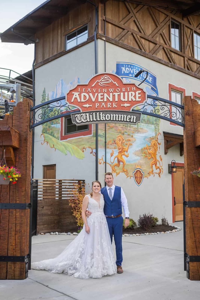 A bride a groom smile at the camera in front of an adventure park before one of the epic things they decided to do during their elopement... ride a rollercoaster!