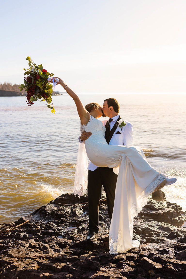 A groom carries his bride across the rocky beach at Gooseberry State Park on their wedding day.