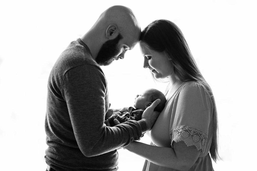 A black and white image shows a mother and father resting their foreheads on one another while they cuddle their baby to their chests during their Twin Cities Lifestyle Newborn Photography session in Minnesota.