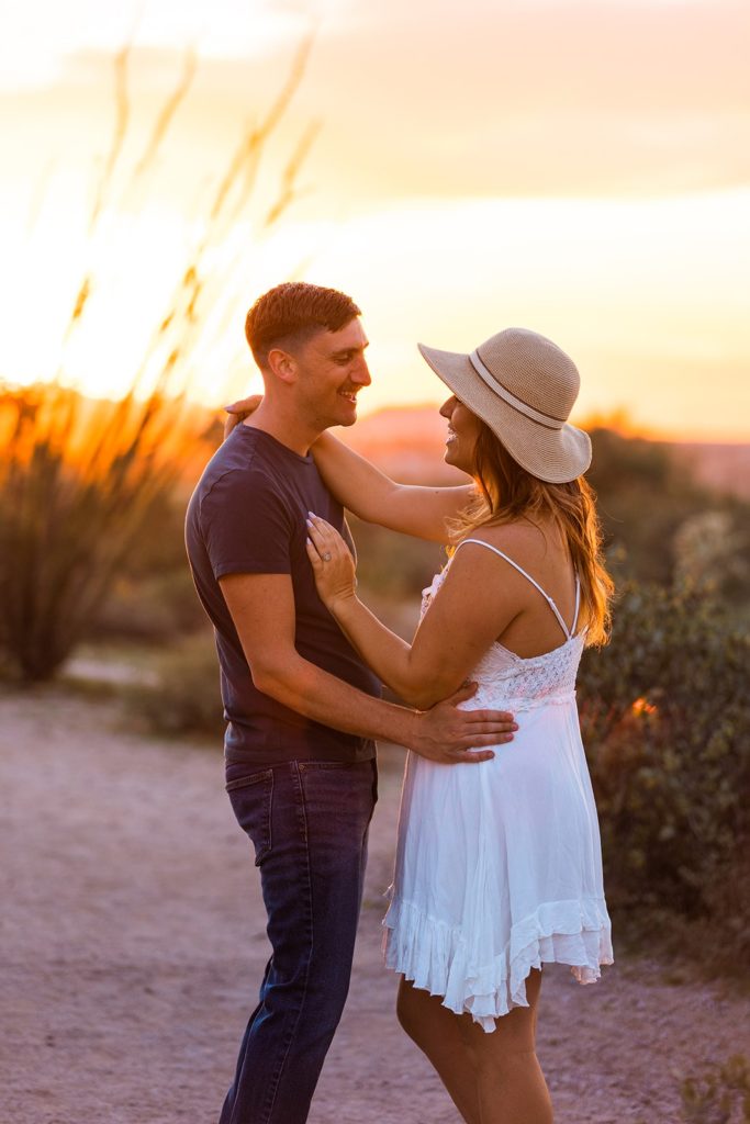 The desert sun glows bright orange as it sets behind a couple embracing during their Lost Dutchman engagement photoshoot in Arizona.