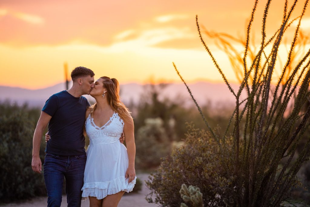 A couple kiss while walking among the cacti at Lost Dutchman State Park during their engagement photoshoot with Forever and Evergreens Photography.
