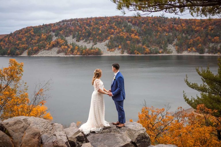 A couple stands on a large rock holding hands and looking out over Devil's Lake during their elopement in the midwest of the United States. Fall colors show brightly in the leaves around them and across the lake.