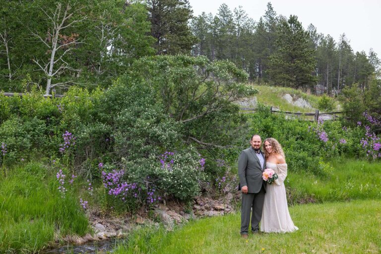 A bride and groom pose for a photo during their Black Hills elopement in South Dakota. Behind them, a hills is covered in purple and white flowers and a wall of pine trees tops the hill. The gorgeous scenery makes it clear that this is one of the most beautiful places to get married in the midwest.