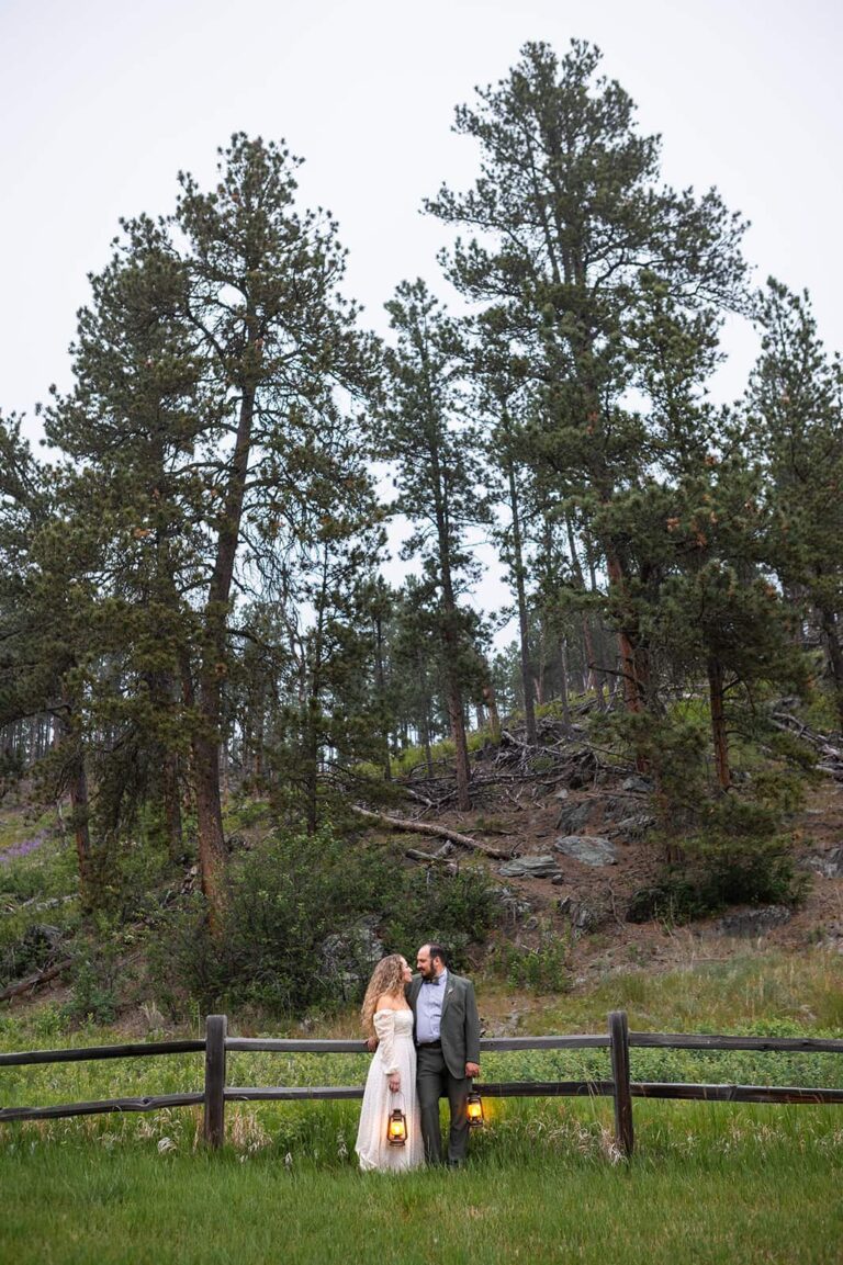 A couple stands in front of an old wooden fence holding glowing lanterns in their wedding attire. Behind them, towering pines grow of a rocky hill. The backdrop is a perfect example of the beauty that the Black Hills of South Dakota has to offer and one of the reasons it is one of the best places to elope in the Midwest.