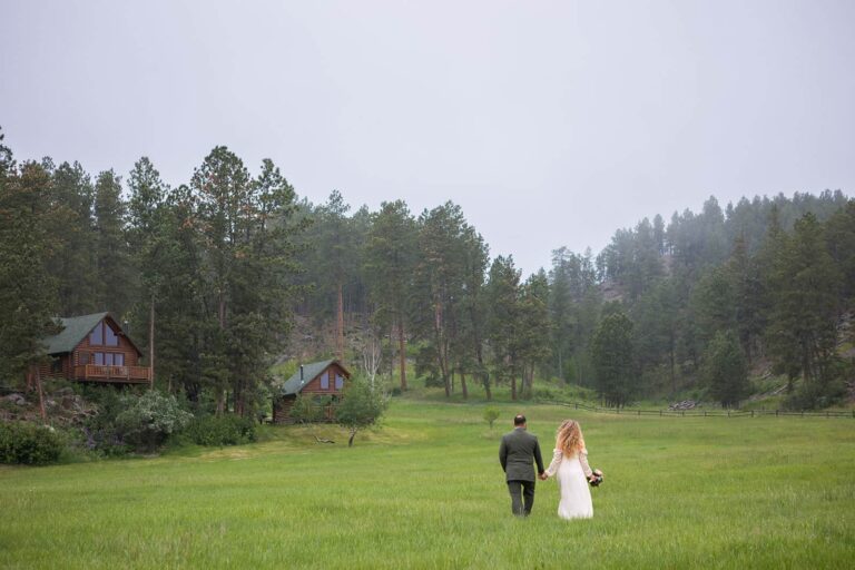 A couple in their wedding clothes walks through a grassy field towards to cabins on a hill during their Black Hills elopement in South Dakota. It's an overcast day and they hold lanterns to light their way.