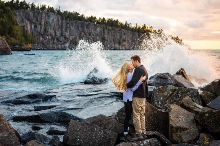 A newly engaged couple kiss on the shore of Tettagouche State Park during their engagement photos as a wave crashes on the rocks of Lake Superior behind them.