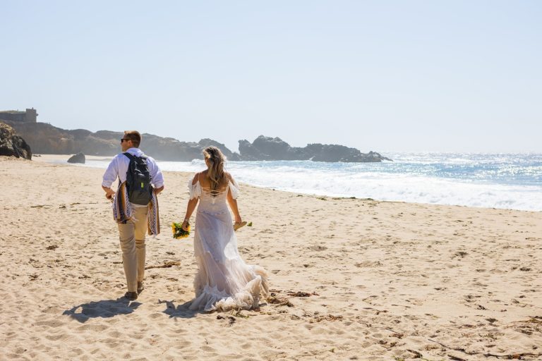 A bride and groom hike along the beach in their wedding clothes in search of the perfect picnic spot during their Big Sur elopement. The bride is hiking in her wedding dress with her train trailing behind her in the sand.