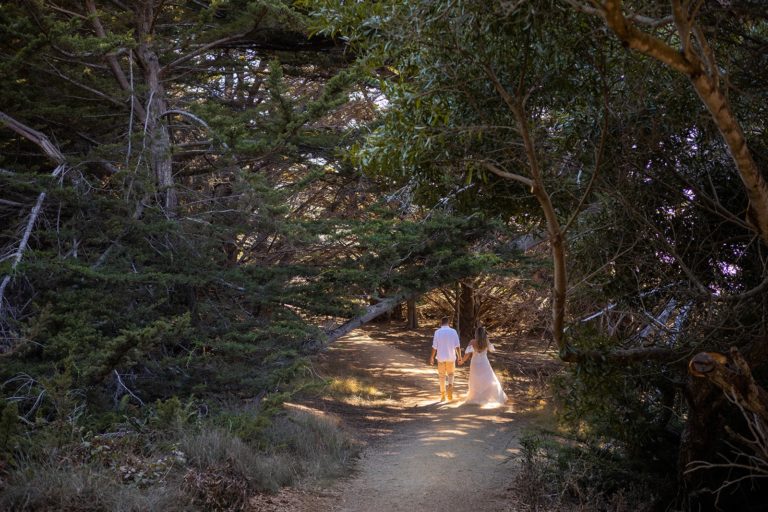 A couple hikes through a tunnel of trees during their Big Sur adventure elopement in California in their wedding clothes. The brides dress kicks up some dust as it drags through the dirt while she is hiking in wedding dress.