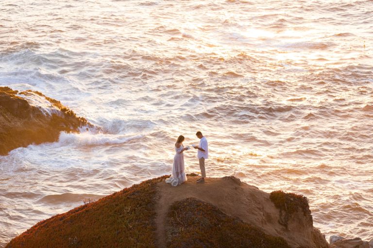 A bride and groom say their vows on their wedding day as the ocean lights up with the colors of the sunset behind them along the California coast.