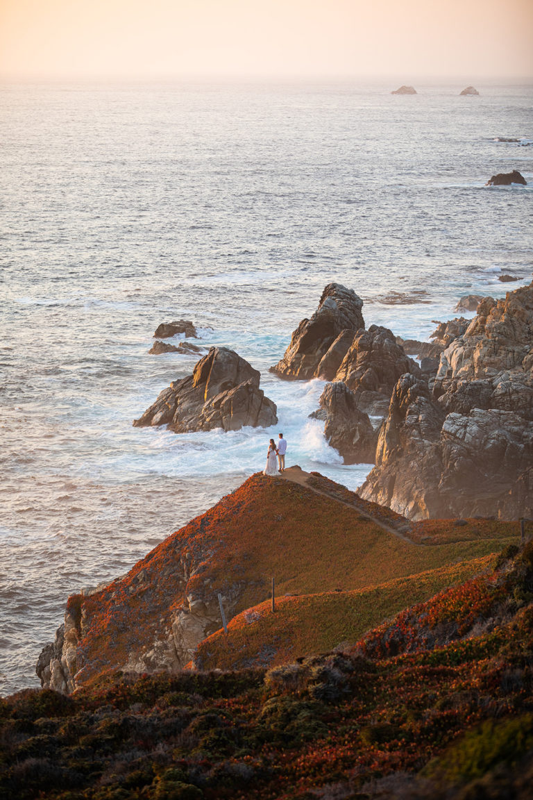 A couple standing on a cliff overlooking the ocean look out at the sunset during their Big Sur elopement.
