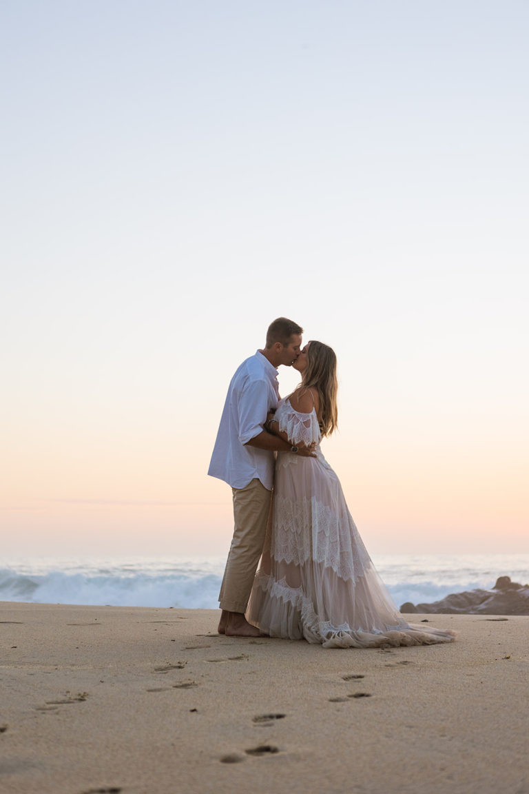 A bride and groom kiss on the beach during their elopement in Big Sur, California. Footprints in the sand indicate they that have been hiking along the beach in their wedding attire. The train of the brides dress is sanding from hiking in her wedding dress.
