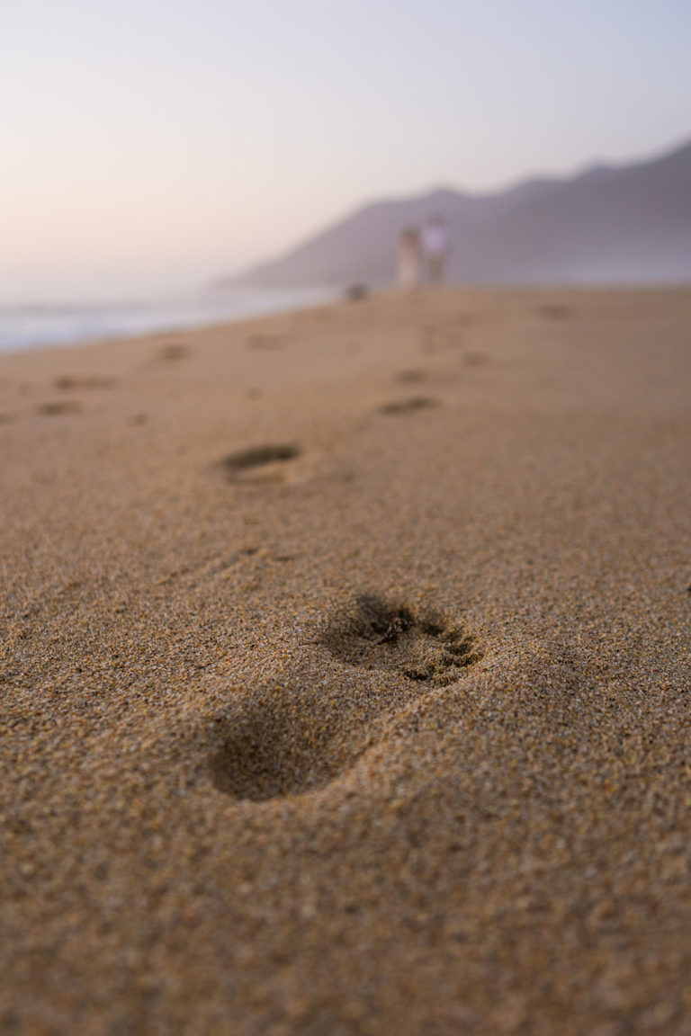 Footprints in the sand lead to a couple walking along the beach of the California coast during their elopement.