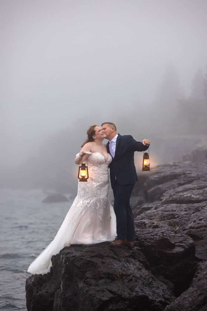 A groom kisses his brides cheek as the both hold lanterns on a foggy morning standing on the rocky shore of Lake Superior during their elopement in the Midwest.