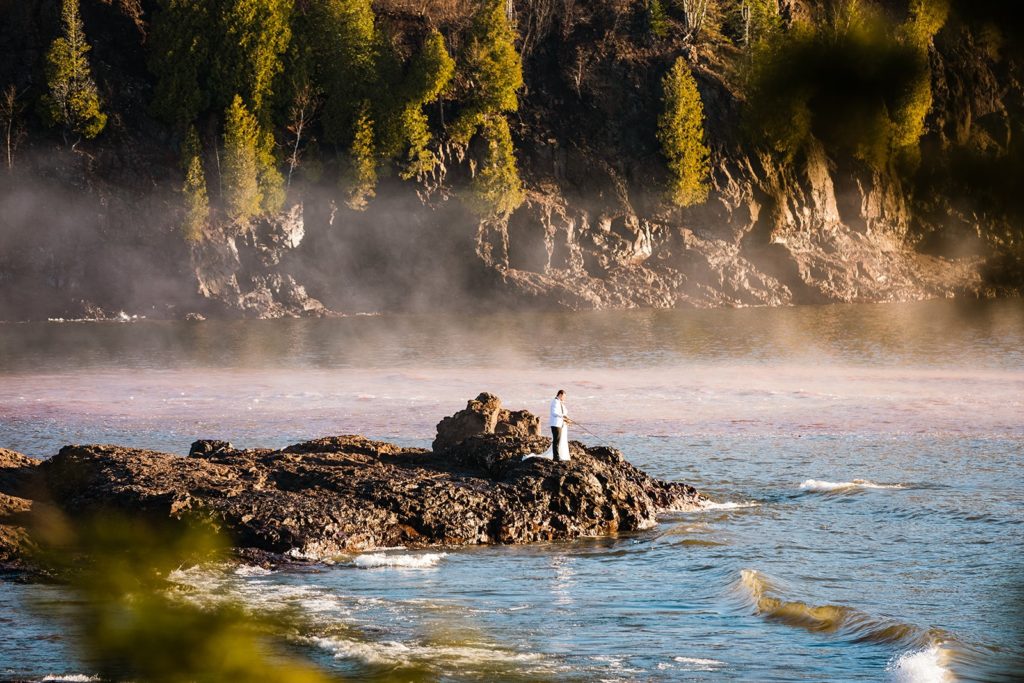 A bride and groom fish in Lake Superior on their wedding day at Gooseberry Falls State Park. Up for adventure, the bride wore her wedding dress while hiking along the rocky shore. Spring is a magical time in Minnesota and one of the best times to elope in Minnesota.