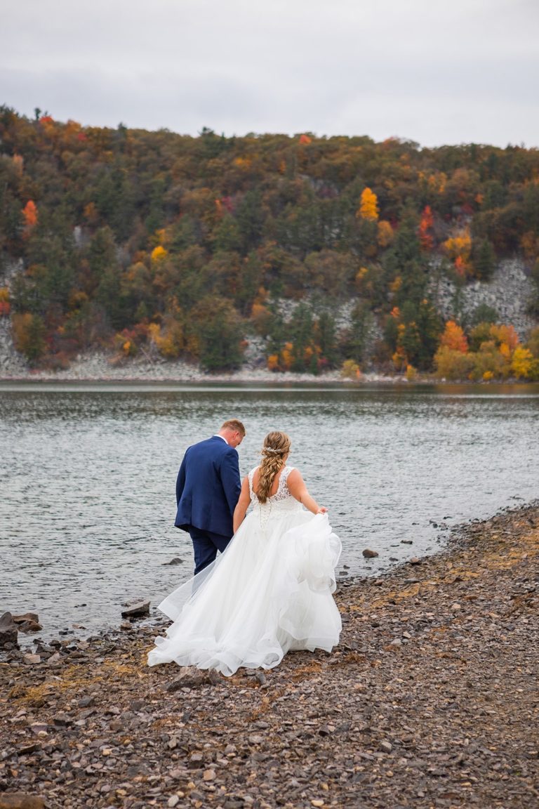 A couple walks along the lake shore in their wedding attire during their fall Devil's Lake elopement.