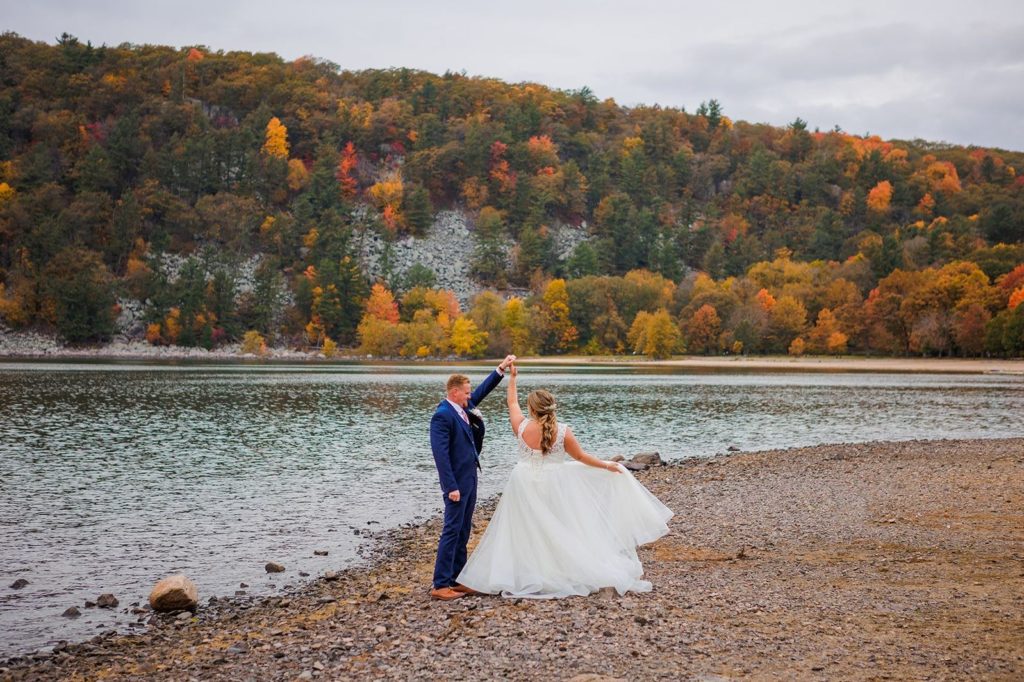 A newly married couple dances along the shore of Devil's Lake during their intimate wedding. The bride twirls her elopement wedding dress as she is spun by her groom. Forever and Evergreens Elopement Photography