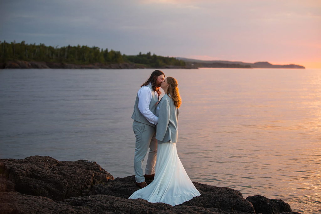 A couple kisses on the North Shore of Lake Superior during their elopement in Minnesota. It's a chilly morning and the brides elopement wedding dress was a bit too thin to keep her warm. The groom has draped his coat over the shoulders of his bride to warm her up.