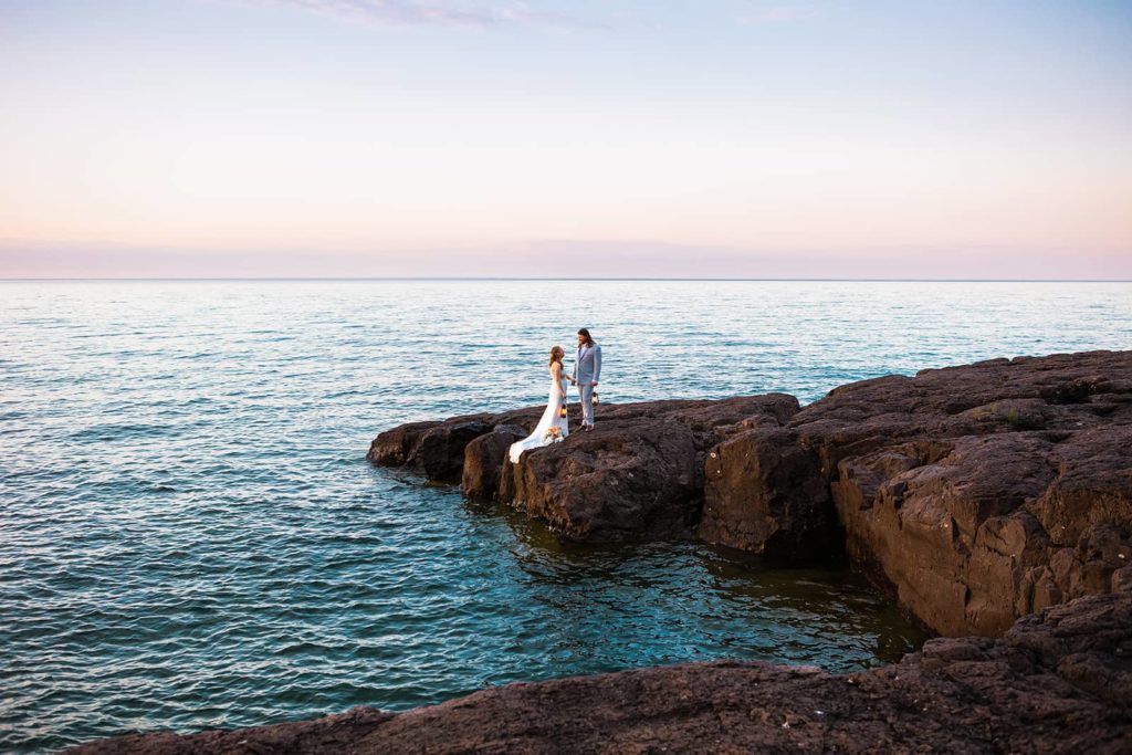 A couple hold hands and grasp their lanterns during sunrise on their elopement day. They stand on a rocky peninsula jutting out into Lake Superior as the first light of day hits the North Shore.