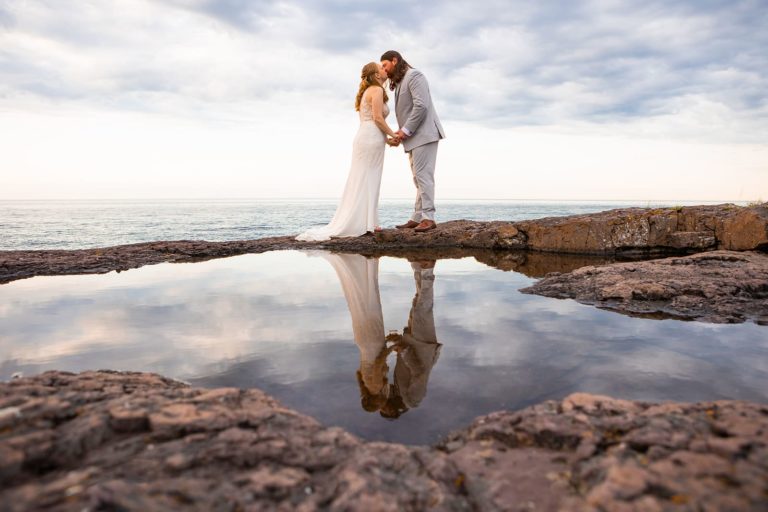 A couple in their wedding clothes hold hands and kiss on the rocky North Shore of Lake Superior in Minnesota during their elopement. In front of them, a large puddle reflects their image perfectly, acting as a large mirror.