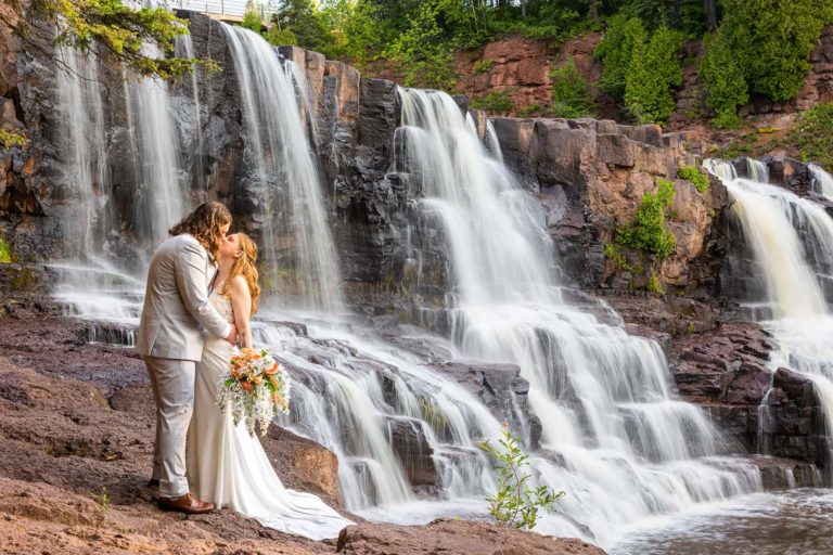 A couple kiss in front of a rushing waterfall during their July North Shore elopement in Minnesota with Forever and Evergreens Photography.