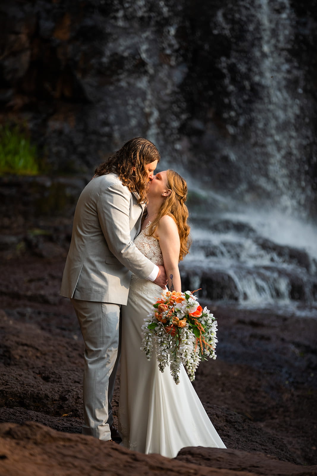 Standing in front of one of the many waterfalls that line the north shore of lake superior, a bride and groom kiss on their elopement day. The bride holds a cascading bouquet of wedding flowers in her hand.