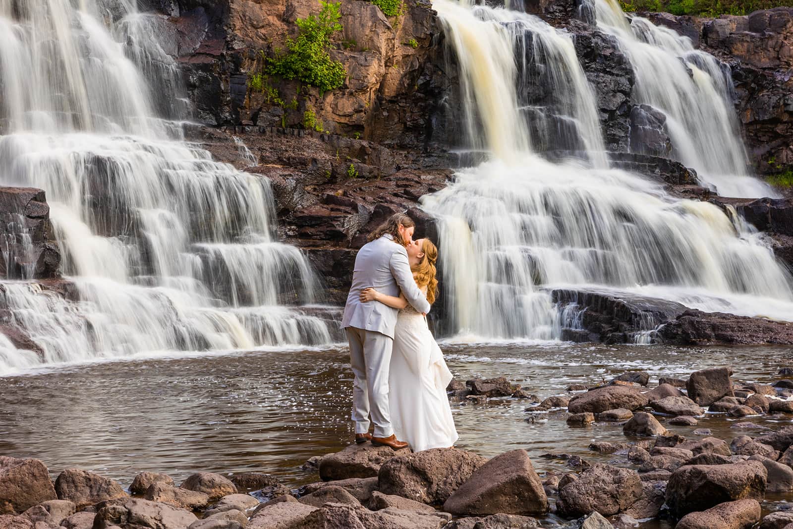 A bride and groom stand on some rocks in the river in front of Gooseberry Falls during their North Shore adventure elopement.