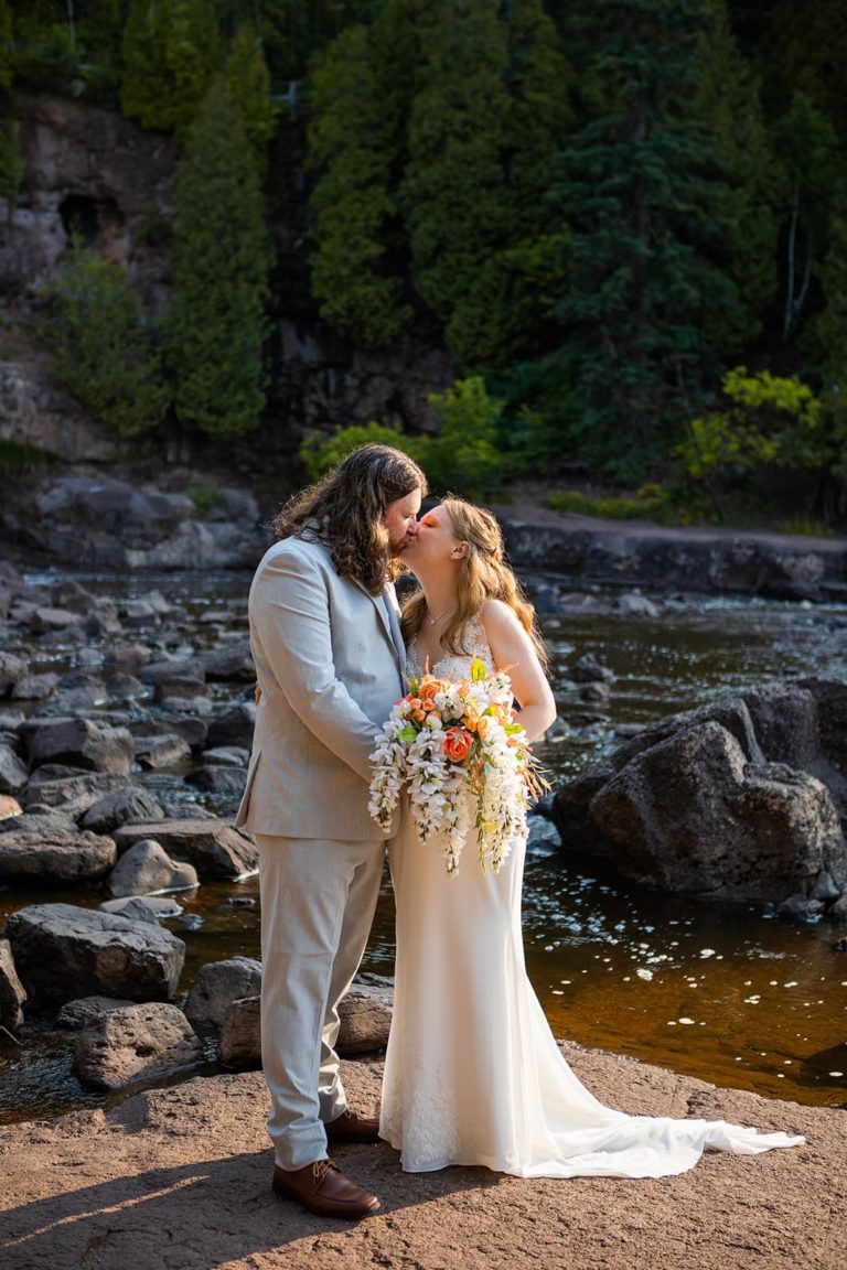 A bride and groom kiss in front of a rocky river during their elopement along the North Shore of Lake Superior.