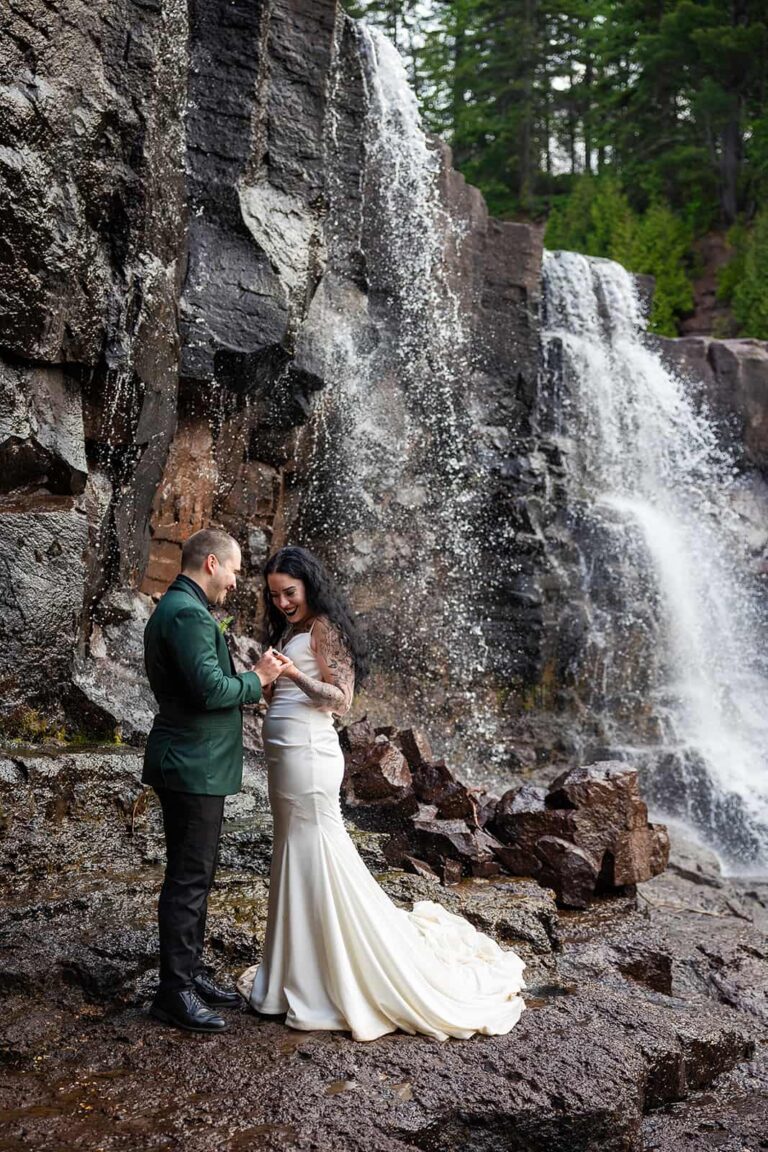 A groom in a green suit and a bride in a flowing white wedding dress stand in front of a waterfall holding hands during their elopement along the Minnesota North Shore.