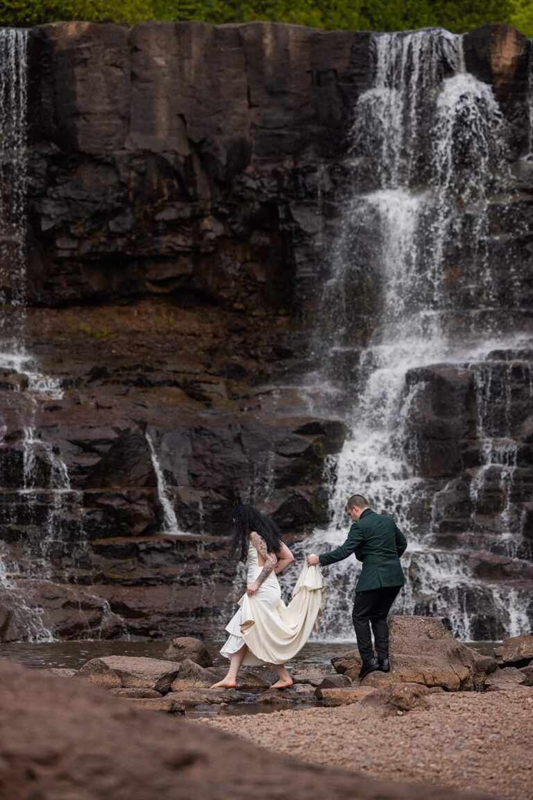 A groom holds the train of his wife's wedding dress and they skip across the rocks of a river during their North Shore wedding in Minnesota.