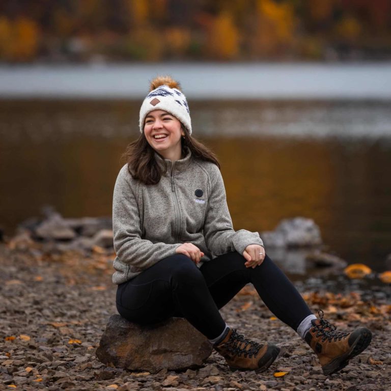 Tali, one of the elopement photographers for Forever and Evergreens, sits on a rock near the edge of Devil's Lake in Wisconsin. It's a brisk fall day in the state park and the fall colors can be seen on the trees across the lake.