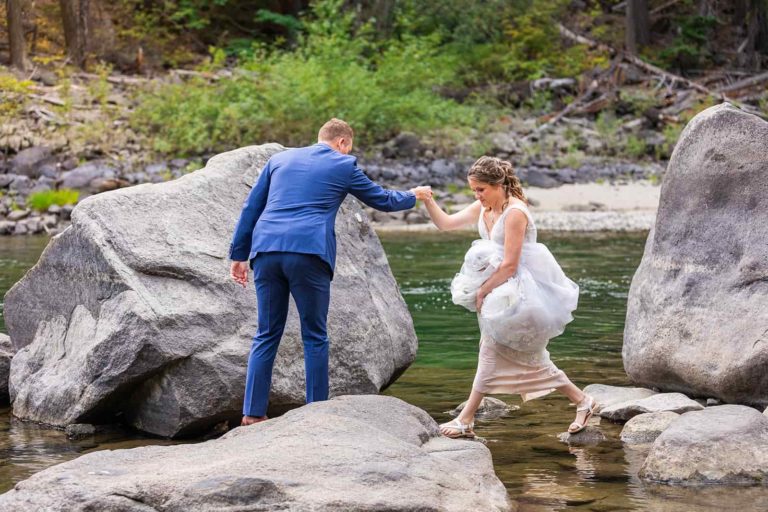 A couple jump across rocks in a river on their elopement day. The bride holds up her elopement wedding dress to keep it out of the water as the groom reaches out his hand to help her across.