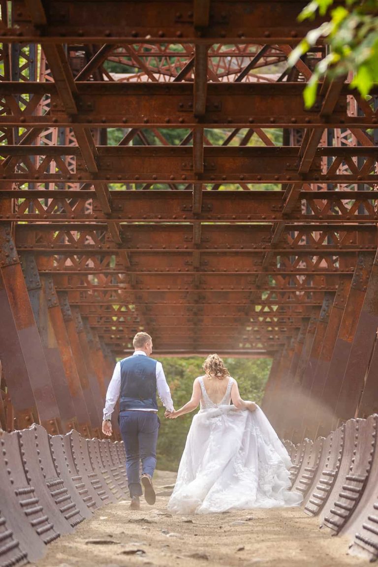 Dust kicks up around their feet as a bride and groom run across a bridge in their wedding clothes during their elopement. The bride's elopement wedding dress blows in the wind as she runs.