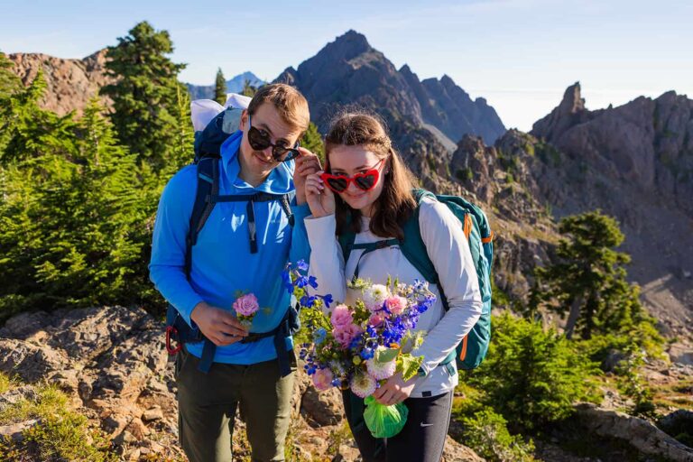 A bride and groom smile over their heart shaped sunglasses on top of a mountain in Washington state. They are wearing hiking clothes with big backpacks. Carrying your wedding dress in a hiking backpack is a great alternative to hike in a wedding dress.