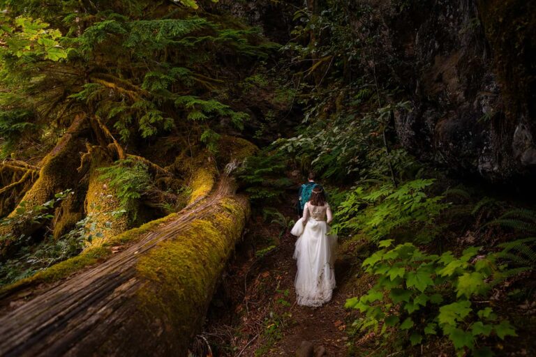 A bridge and groom hike through this thick forests of Washington during their adventure elopement. The train of the bride's wedding dress drags through the dirt as she hikes in her wedding dress.