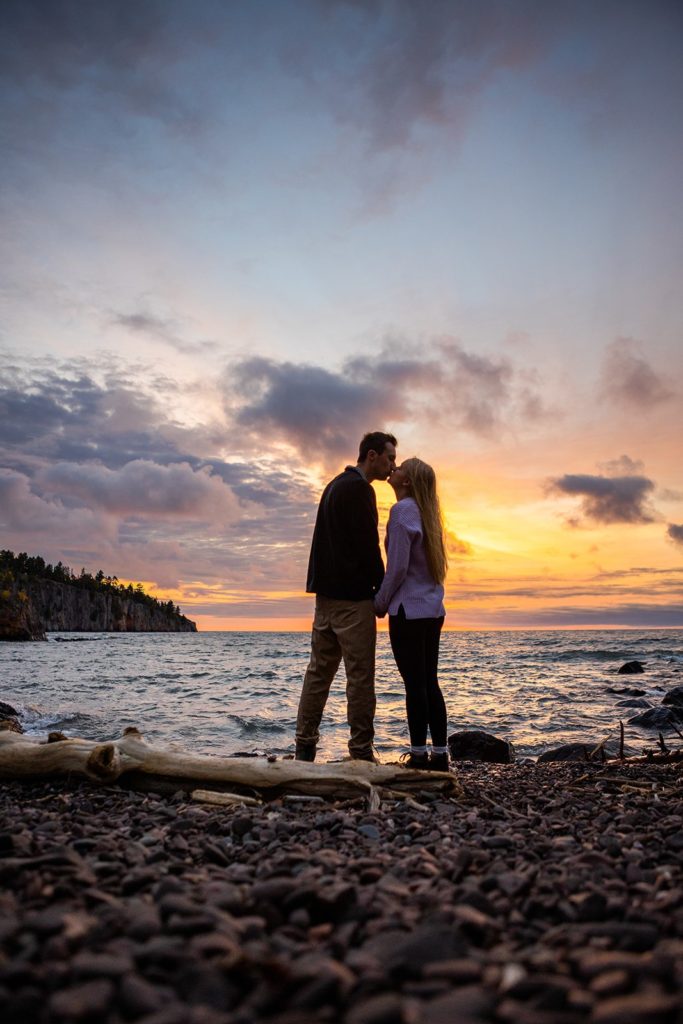 A couple poses on the beach overlooking a colorful sunrise over Lake Superior during their Tettegouche State Park engagement photos.