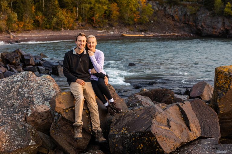 A couple smiles at the camera in the warm light of sunrise while they sit on a jagged rock during their Tettegouche State Park engagement photo shoot.