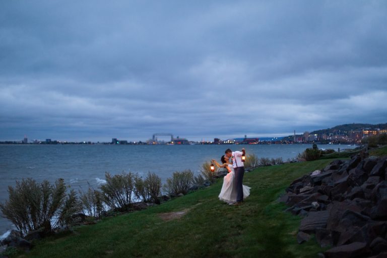 A couple embrace illuminated by the glow of their lanterns along the shore of Lake Superior on a moody fall evening. The Duluth Lift Bridge can be seen along the skyline behind them. Beautiful weather is one of the reasons we think fall is the best time of year to elope in Minnesota!