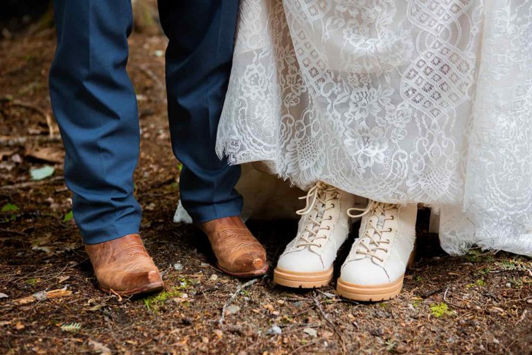A bride hikes up her elopement wedding dress to show off her hiking boot. Her groom stands next to her in cowboy boots. Proper footwear is important while hiking on your elopement day.