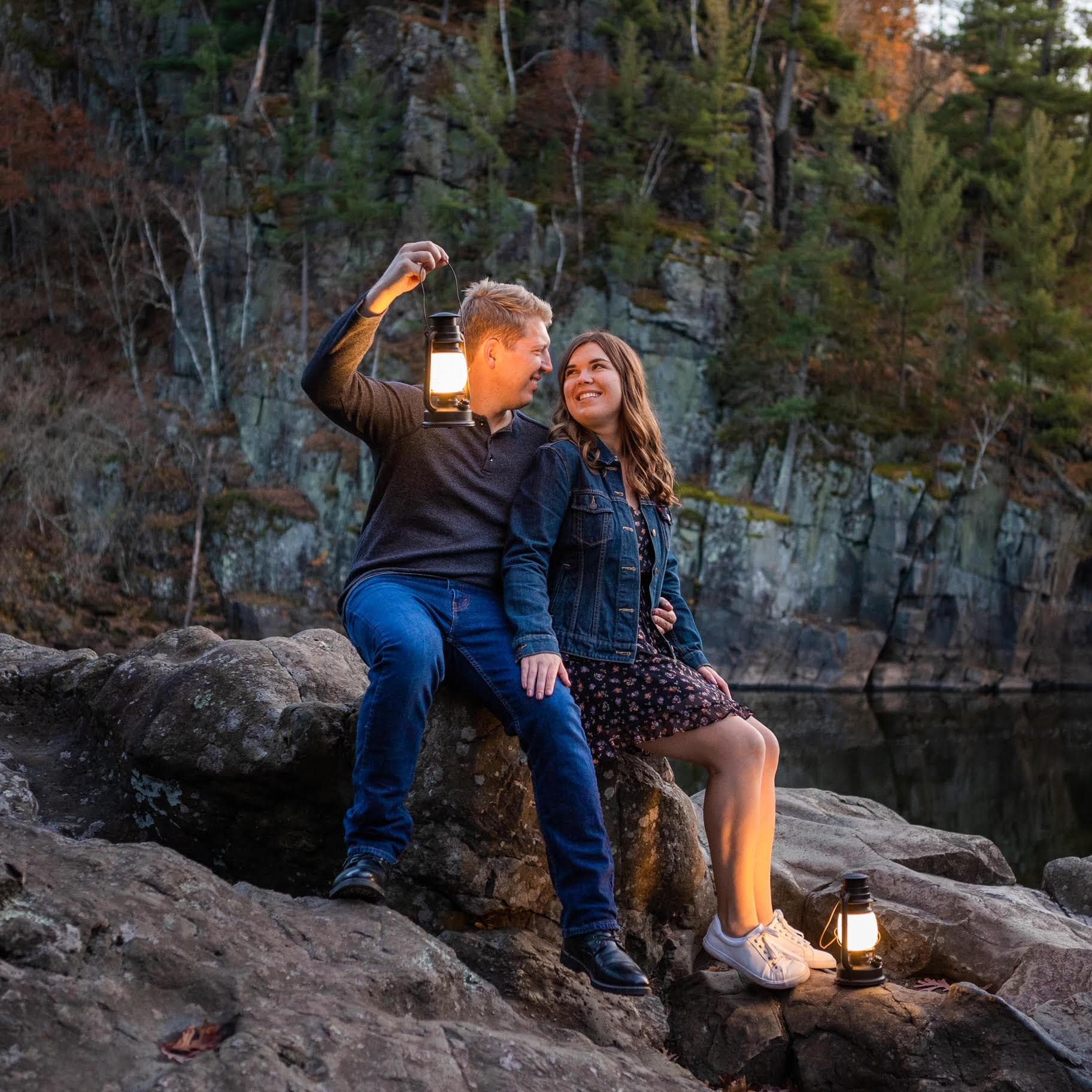 A couple sits along a rocky river holding lanterns while having their engagement photos taken by Minnesota engagement and elopement photographers Forever and Evergreens.