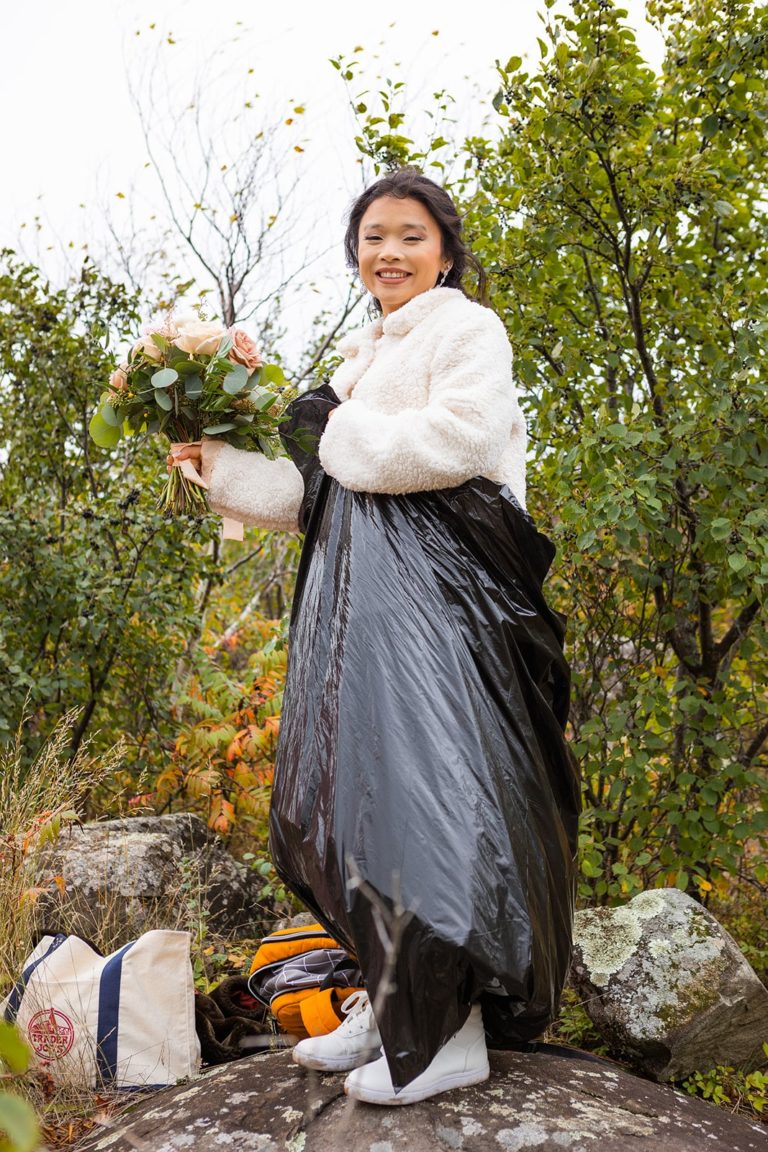 A bride smiles at the camera while wearing a cute white jacket and a black trash bag. Her wedding dress is tucked into the trash bag to keep it clean while she hike in wedding dress during her fall adventure elopement along the North Shore of Lake Superior in Duluth, MN.